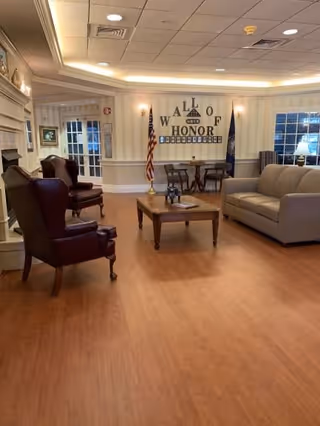 A cozy living room area with wooden flooring, two dark leather armchairs on the left, a beige sofa on the right, and a wooden coffee table in the center. The back wall features a display titled 'Wall of Honor' with an American flag and another flag on either side. The room has soft lighting and large windows.
