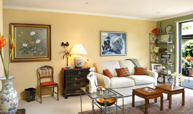 Sunlit living room with a white sofa, coffee tables, framed artwork on yellow walls, and an open doorway to a patio.