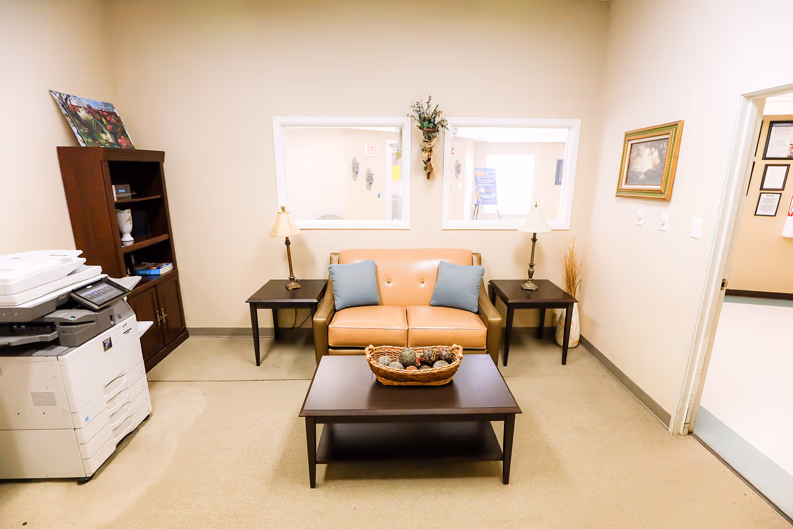 A small waiting or sitting area with a tan leather loveseat with two blue pillows, flanked by two dark wooden side tables each with a lamp. In front of the loveseat is a dark wooden coffee table with a decorative basket. To the left is a dark wooden bookshelf and a large white office printer. The walls are beige with two windows looking into another room, and a framed picture hangs on the right wall.