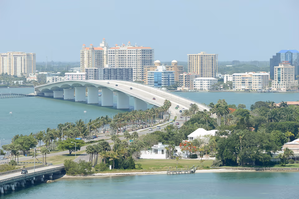 A scenic view of a large bridge spanning over a body of water with several cars traveling on it. The bridge connects to a cityscape with multiple tall buildings and palm trees lining the waterfront. The foreground features lush greenery and a few houses near the water's edge under a clear sky.