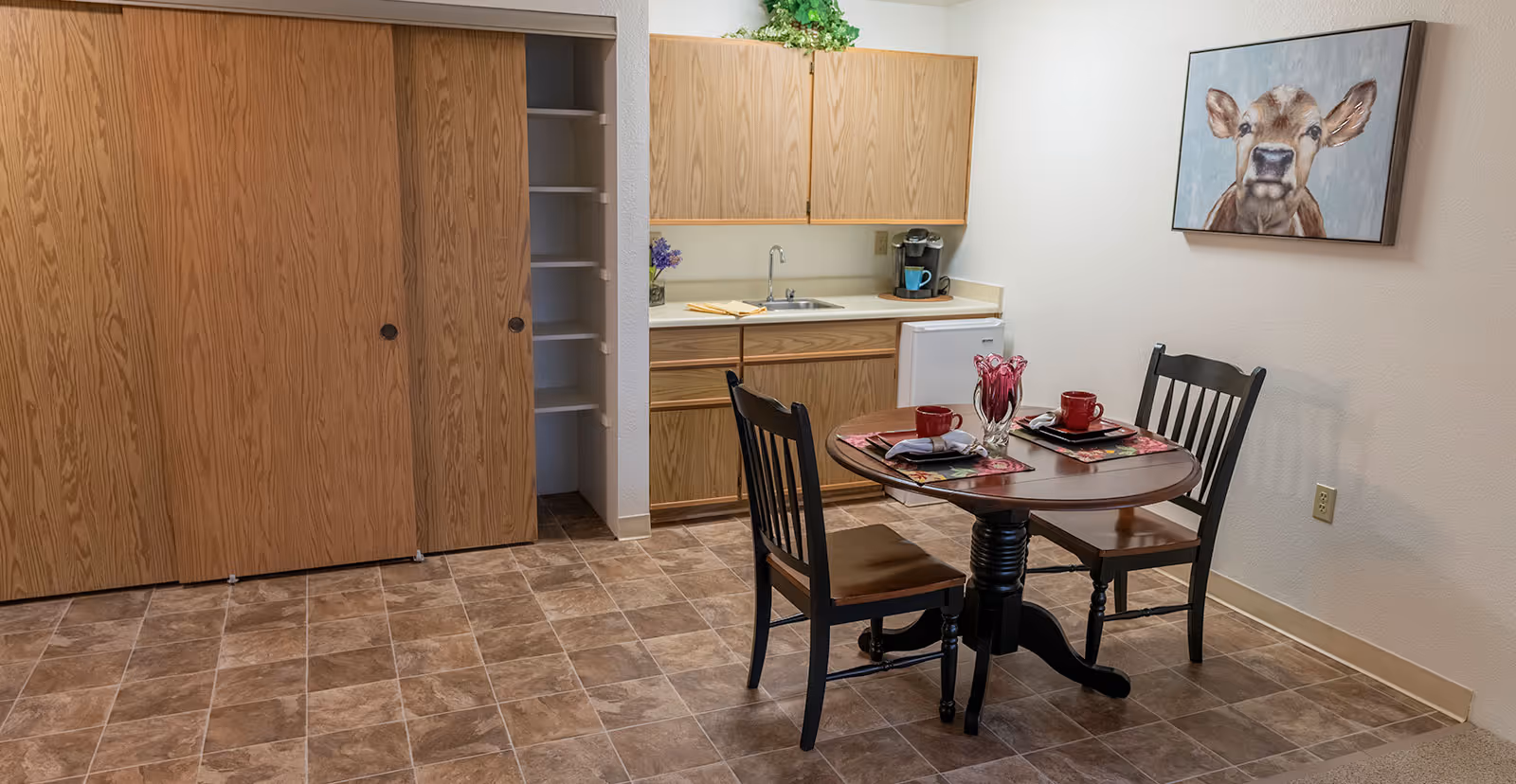 A small dining area in a senior living facility with a round wooden table set for two with red cups and floral placemats. Two wooden chairs are placed around the table. Behind the table is a kitchenette with wooden cabinets, a small sink, a coffee maker, and a mini refrigerator. To the left, there is a large wooden closet with sliding doors, one of which is partially open revealing shelves. A painting of a calf hangs on the wall above the dining table.