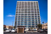 Tall multi-story residential building with balconies on each floor under a clear blue sky, with a parking lot and several parked cars in the foreground.