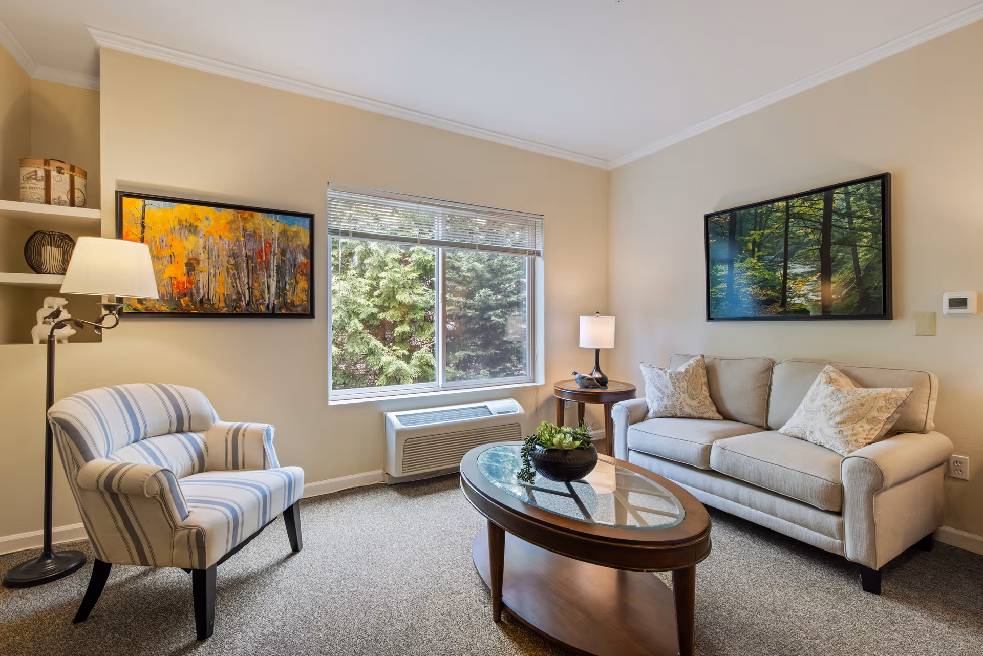A cozy living room with a beige sofa adorned with two patterned pillows, a striped armchair, a wooden oval coffee table with a glass top and a small plant centerpiece, a floor lamp, and a side table with a lamp. The room has beige walls with two framed paintings and a window showing green trees outside.