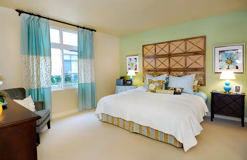 A bright and cozy bedroom with a large bed featuring multiple decorative pillows and a white bedspread. The bed has a wooden headboard with a geometric pattern. On either side of the bed are nightstands with lamps and framed floral artwork above them. A window with blue and white curtains lets in natural light. There is a gray armchair and a wooden dresser in the room.