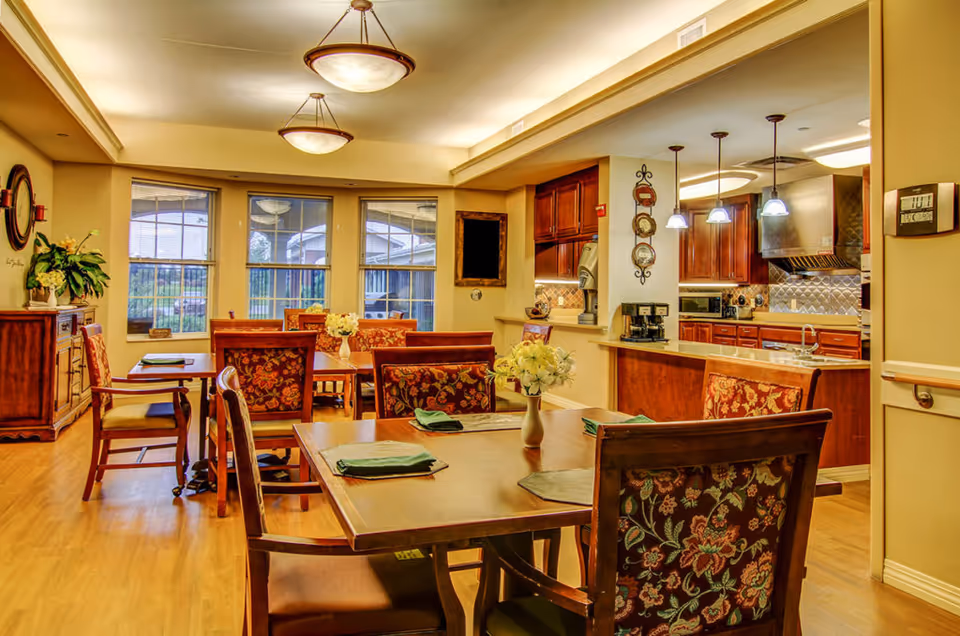 A warm and inviting dining room area with wooden tables and chairs featuring floral upholstery. The tables are set with green napkins and small vases of white flowers. Large windows let in natural light and offer a view of the outdoor area. To the right, there is an open kitchen with wooden cabinets, stainless steel appliances, and pendant lighting above the counter.