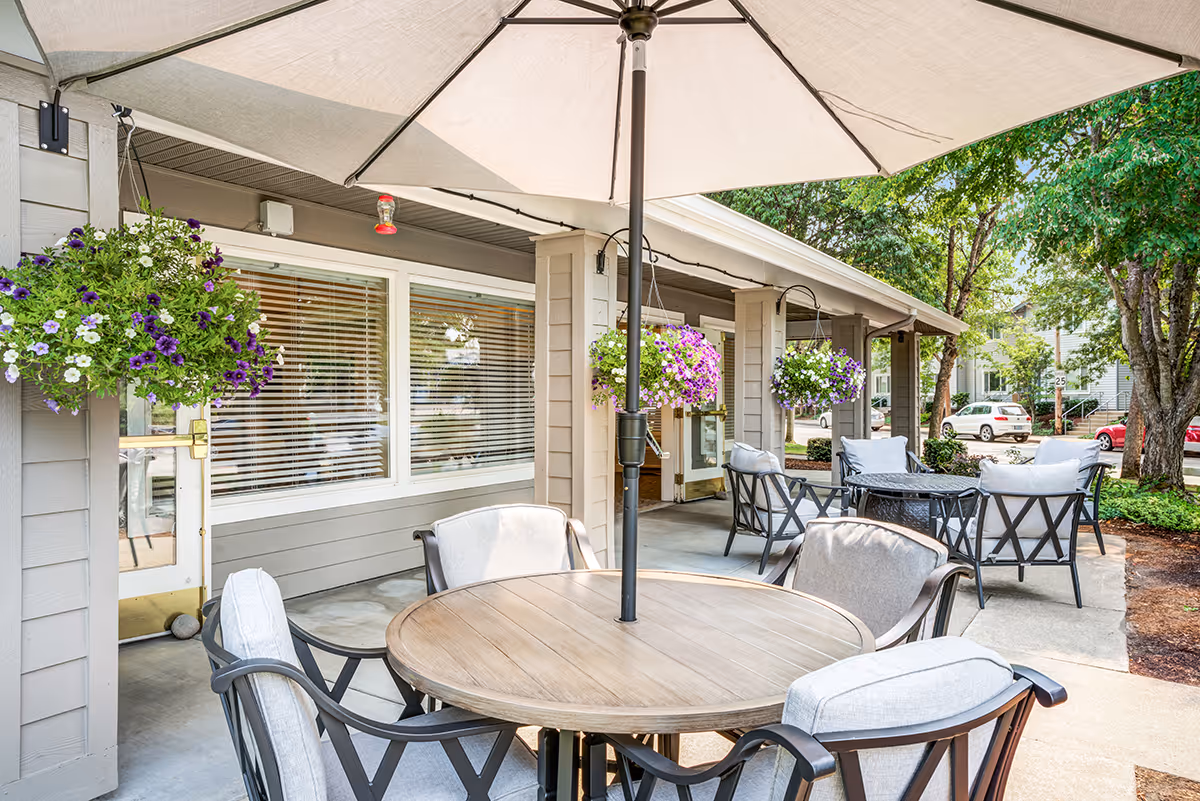 Outdoor patio area at Woodside Senior Living with a round wooden table and cushioned chairs under a large beige umbrella. Hanging flower baskets with purple and white flowers are attached to the building. Additional seating with cushioned chairs and a small table is visible near the entrance. Trees and parked cars are seen in the background.