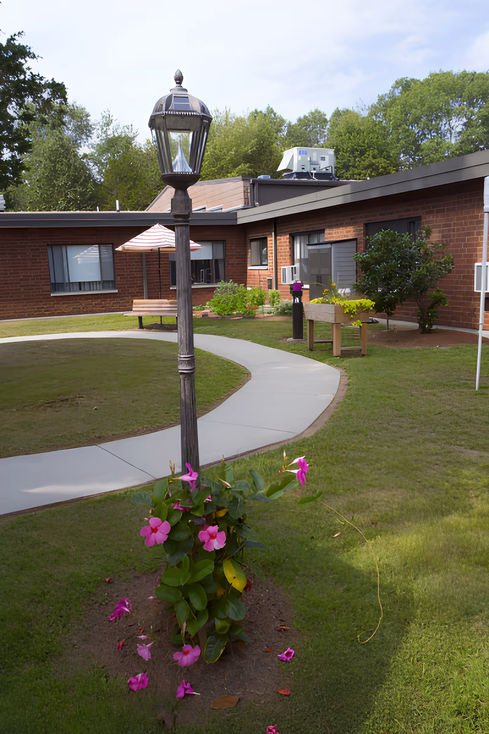 Outdoor courtyard area at Bayberry Commons featuring a curved concrete walkway, a vintage-style lamp post with pink flowers growing at its base, green grass, and a brick building with windows and plants in the background.