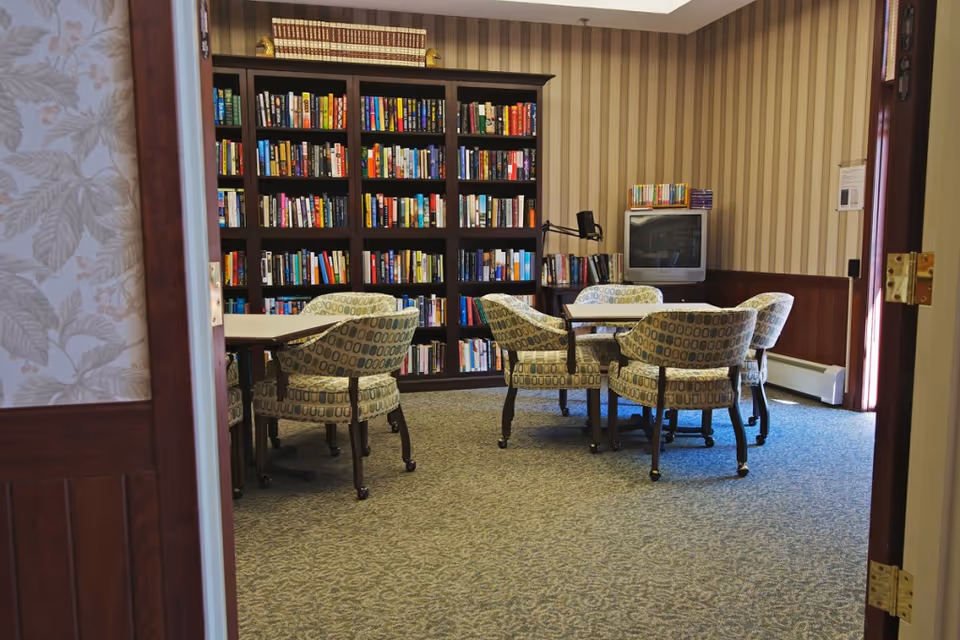 A cozy reading or activity room with patterned armchairs arranged around tables, a large bookshelf filled with books, and a television in the corner. The room has striped wallpaper and carpeted flooring.