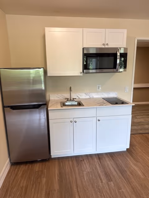 Small kitchen area with a stainless steel refrigerator on the left, white cabinets above and below a marble countertop. The countertop includes a small sink and an electric cooktop. Above the cooktop is a stainless steel microwave. The floor is wood, and there is a doorway to another room on the right.