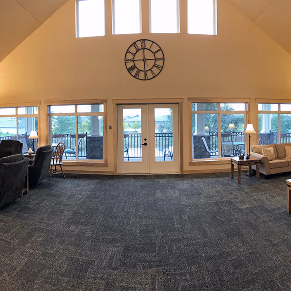 Spacious common lounge with sofas and chairs, large windows and double doors beneath a decorative wall clock.