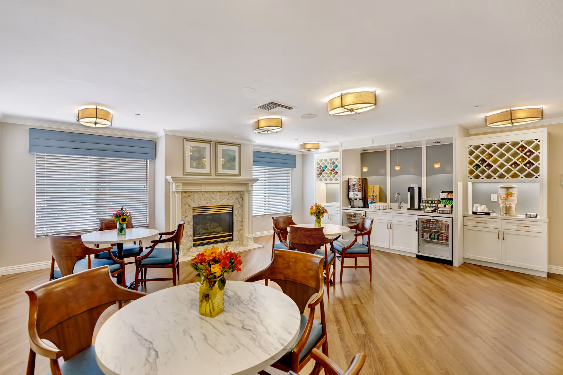 A bright and inviting dining area with round marble-top tables and wooden chairs with blue cushions. The room features a fireplace with a decorative mantle and framed artwork above it. There are two large windows with blue valances, and a beverage station with coffee machines, a water dispenser, and a mini fridge. The floor is wood, and the ceiling has multiple modern light fixtures.