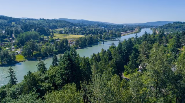 A scenic aerial view of a river winding through a lush green forested area with hills in the background under a clear blue sky.