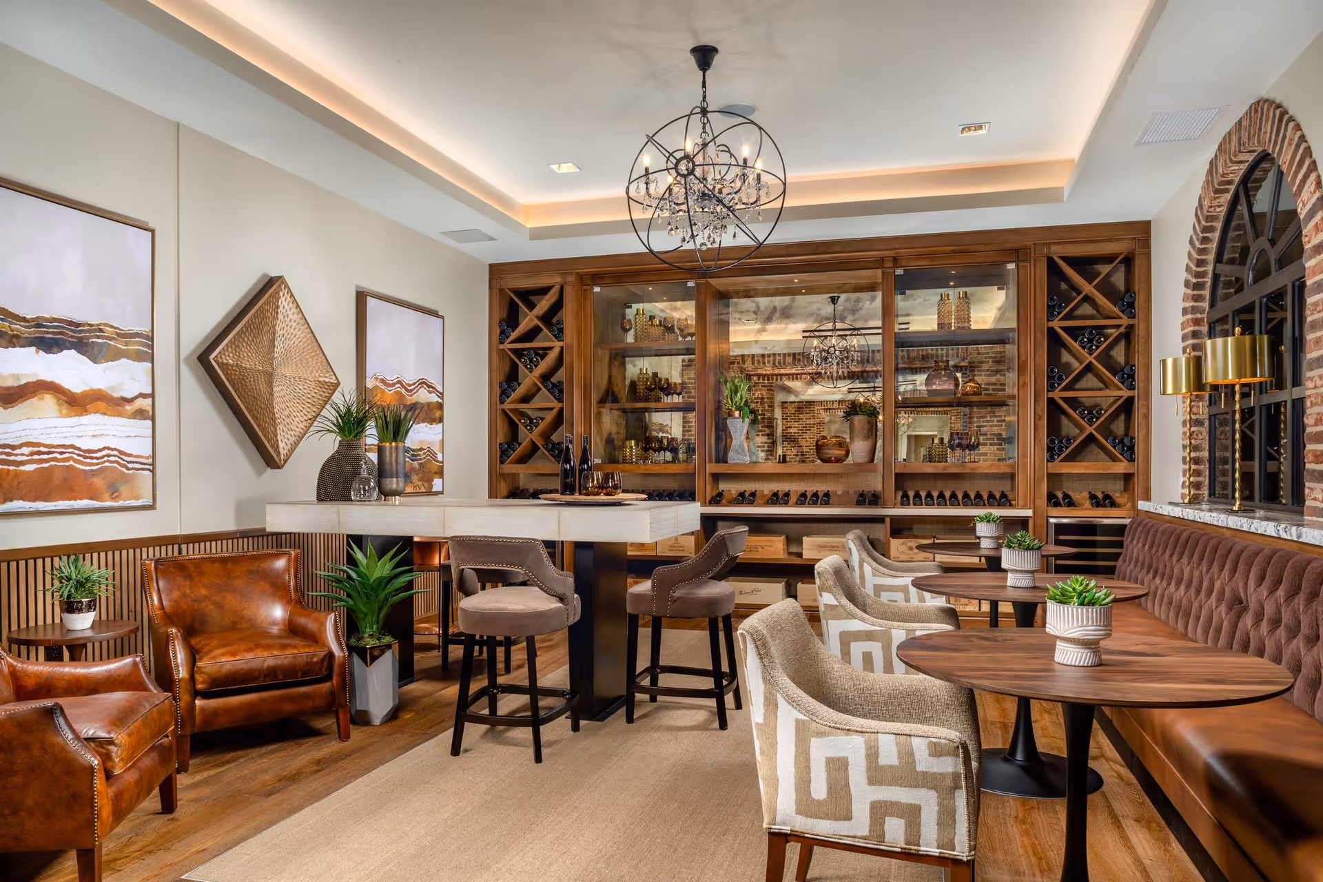 A stylish lounge area with a mix of leather and upholstered chairs around wooden tables. The room features a large wooden wine cabinet with glass doors filled with bottles and decorative items. There are modern light fixtures hanging from the ceiling, abstract wall art, and several potted plants adding greenery. The space has warm lighting and a cozy, inviting atmosphere.