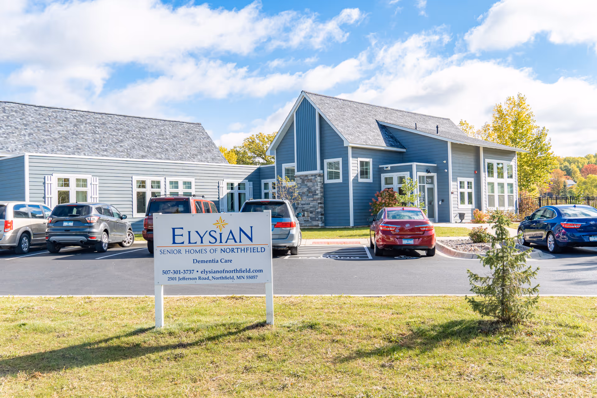 Front exterior of a blue Elysian Senior Homes of Northfield building with parked cars and a sign in the foreground.