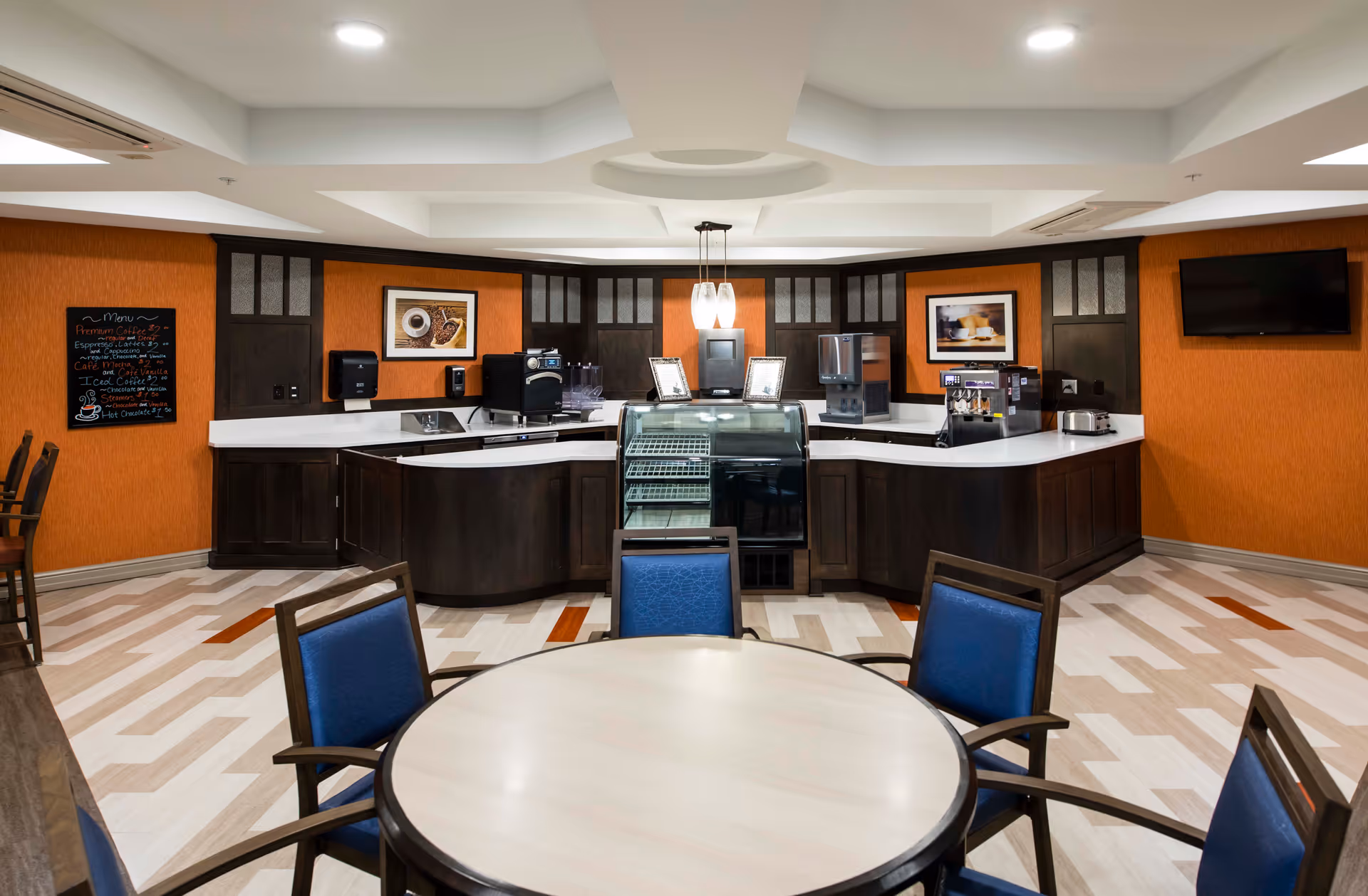 A modern coffee and snack station area in a senior living facility with dark wood cabinetry, white countertops, and orange walls. The space includes a small refrigerated display case, coffee machines, a toaster, and a menu board on the left wall. In the foreground, there is a round table with blue cushioned chairs. A flat-screen TV is mounted on the right wall.
