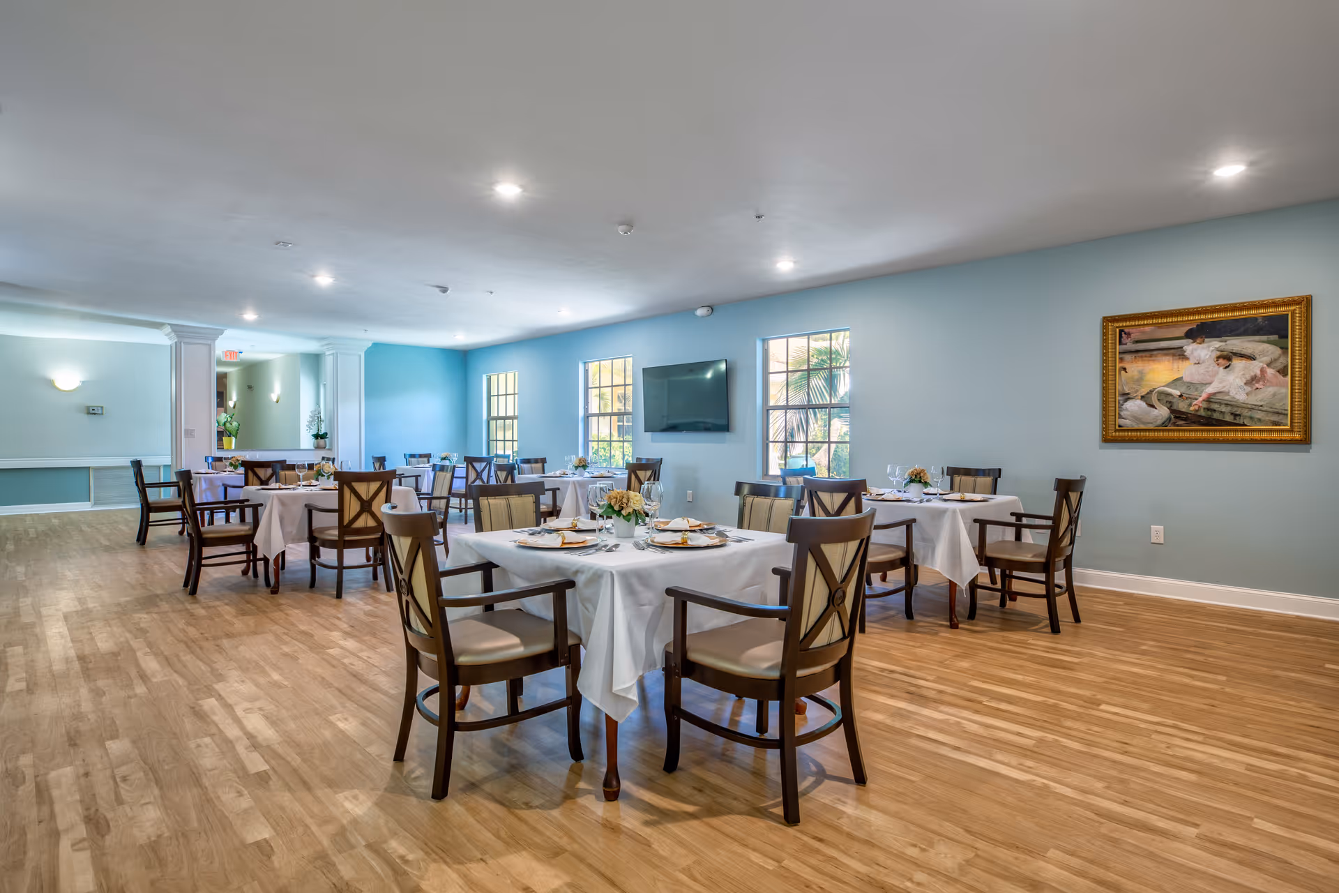 A spacious dining room with several tables covered in white tablecloths, each set with plates, glasses, and floral centerpieces. The room has light blue walls, wooden flooring, and multiple windows letting in natural light. A flat-screen TV is mounted on one wall, and a framed painting hangs on another.