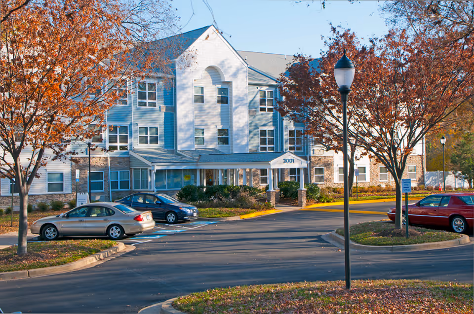 Exterior view of a three-story senior living facility named Rainier Manor, surrounded by autumn trees with orange leaves. There are several parked cars in the parking lot in front of the building, and a streetlamp is visible in the foreground.