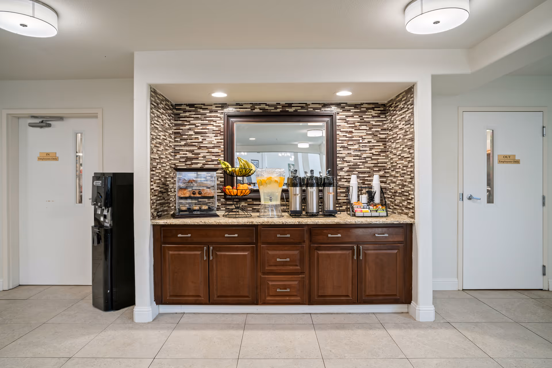 A refreshment station in a hallway with a countertop featuring a fruit basket with bananas and oranges, a container with donuts, a large glass dispenser with lemon water, several coffee thermoses, disposable cups, and a variety of coffee condiments. The backsplash has a mosaic tile design, and there is a large mirror above the counter. On either side of the station are white doors labeled 'IN Employees Only' and 'OUT Employees Only'. A black water cooler is positioned next to the left door.