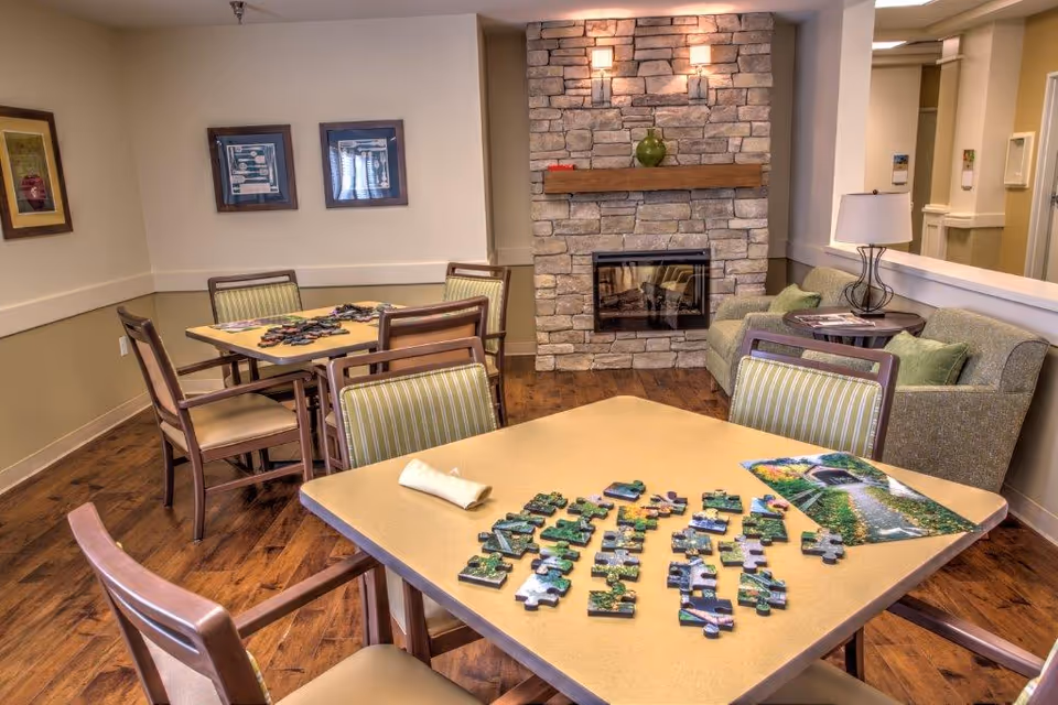 A cozy common area with two tables and chairs, one table has an unfinished jigsaw puzzle and a puzzle box on it. The room features a stone fireplace with a wooden mantle, a green vase on the mantle, a green couch with green pillows, a side table with a lamp, and framed artwork on the walls. The floor is wooden and the walls are painted beige and light brown.