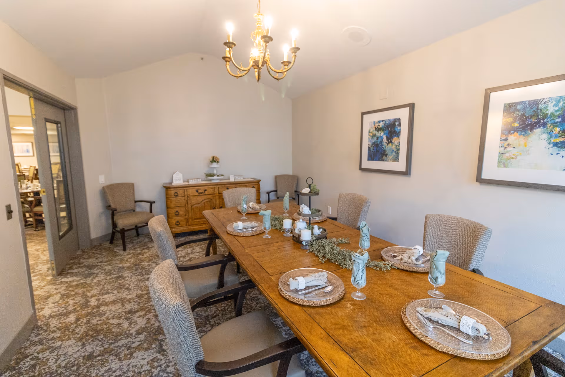 A dining room with a wooden table set for six people, featuring decorative plates, napkins, and glasses. The room has upholstered chairs, a chandelier hanging from the ceiling, two framed abstract paintings on the wall, and a wooden sideboard with decorative items against the far wall.