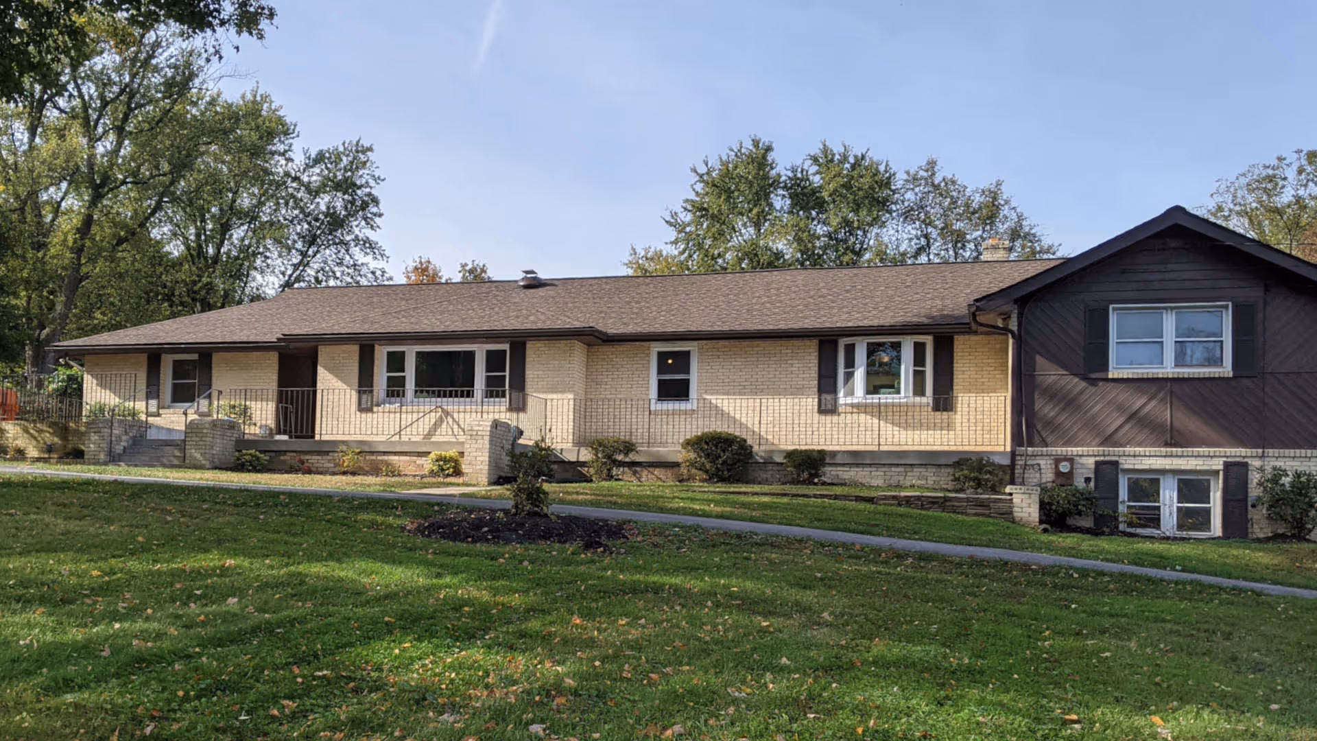 Single-story residential building with beige brick exterior and brown roof, surrounded by green lawn and trees under a clear blue sky.