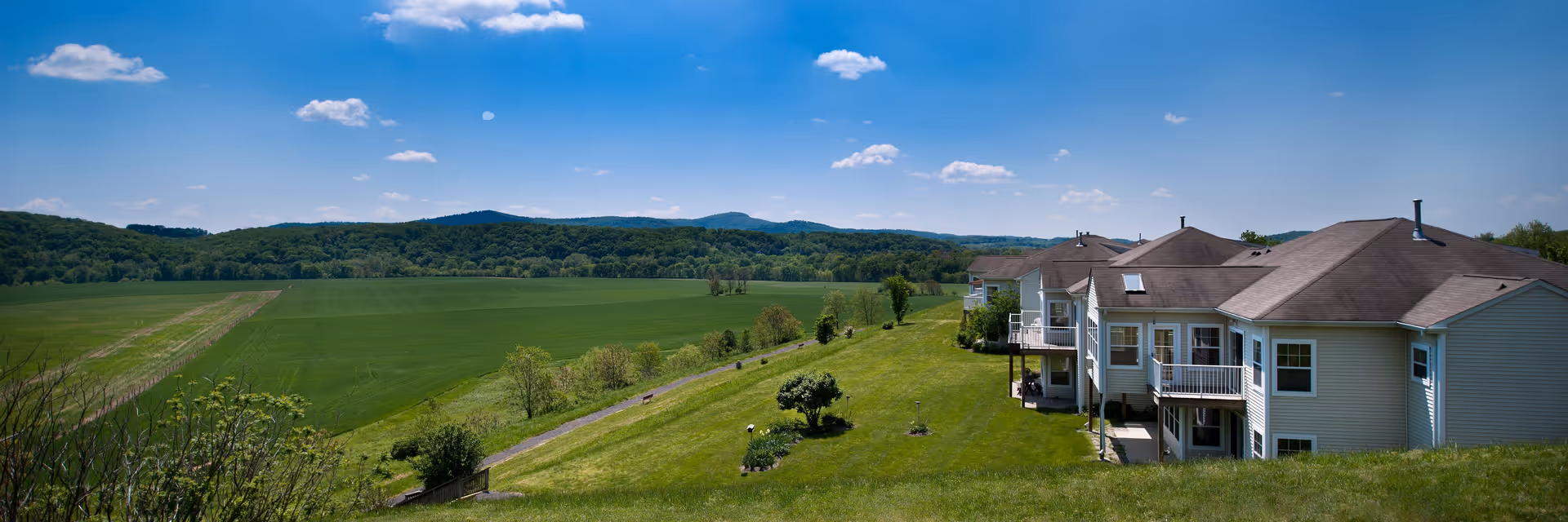 A panoramic view of a senior living facility named Buckingham's Choice with multiple connected buildings featuring balconies, situated on a grassy hill overlooking expansive green fields and forested hills under a bright blue sky with scattered clouds.