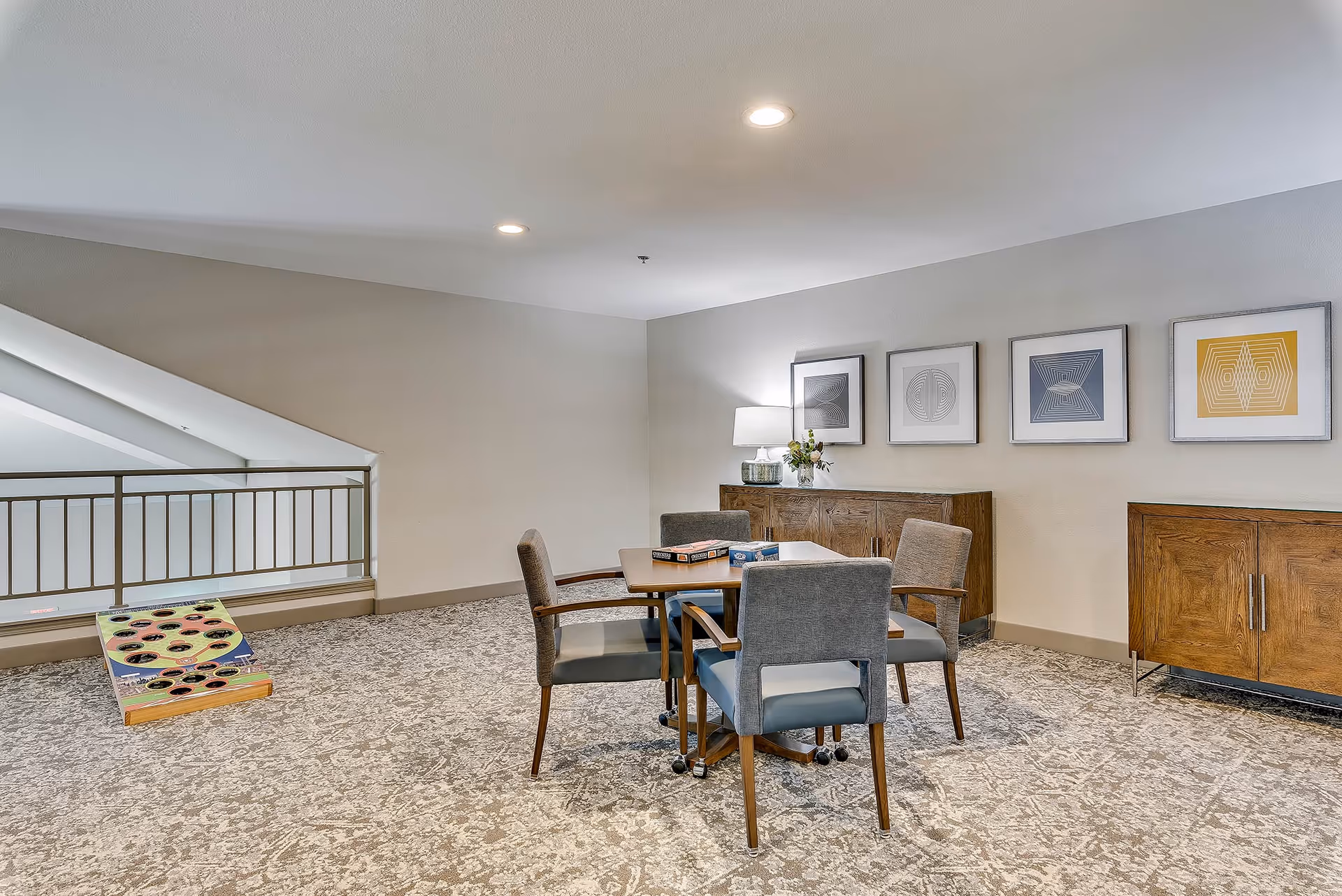 A bright communal seating area with a table and four chairs, wooden cabinets, framed wall art, and a cornhole board near a railing.