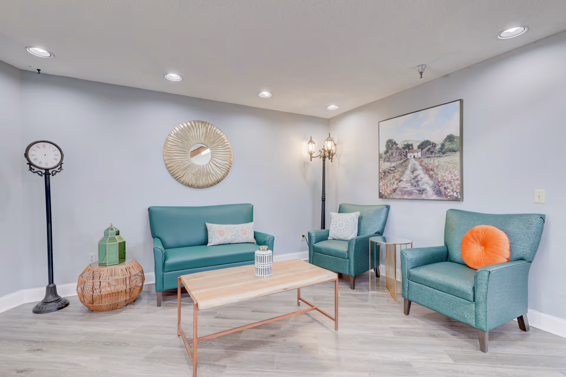 A cozy sitting area with teal upholstered chairs and a loveseat, a wooden coffee table, a round side table, a tall vintage-style clock, a decorative lantern on a wicker basket, a floor lamp with three lights, a round sunburst mirror, and a painting of a countryside path on the wall.