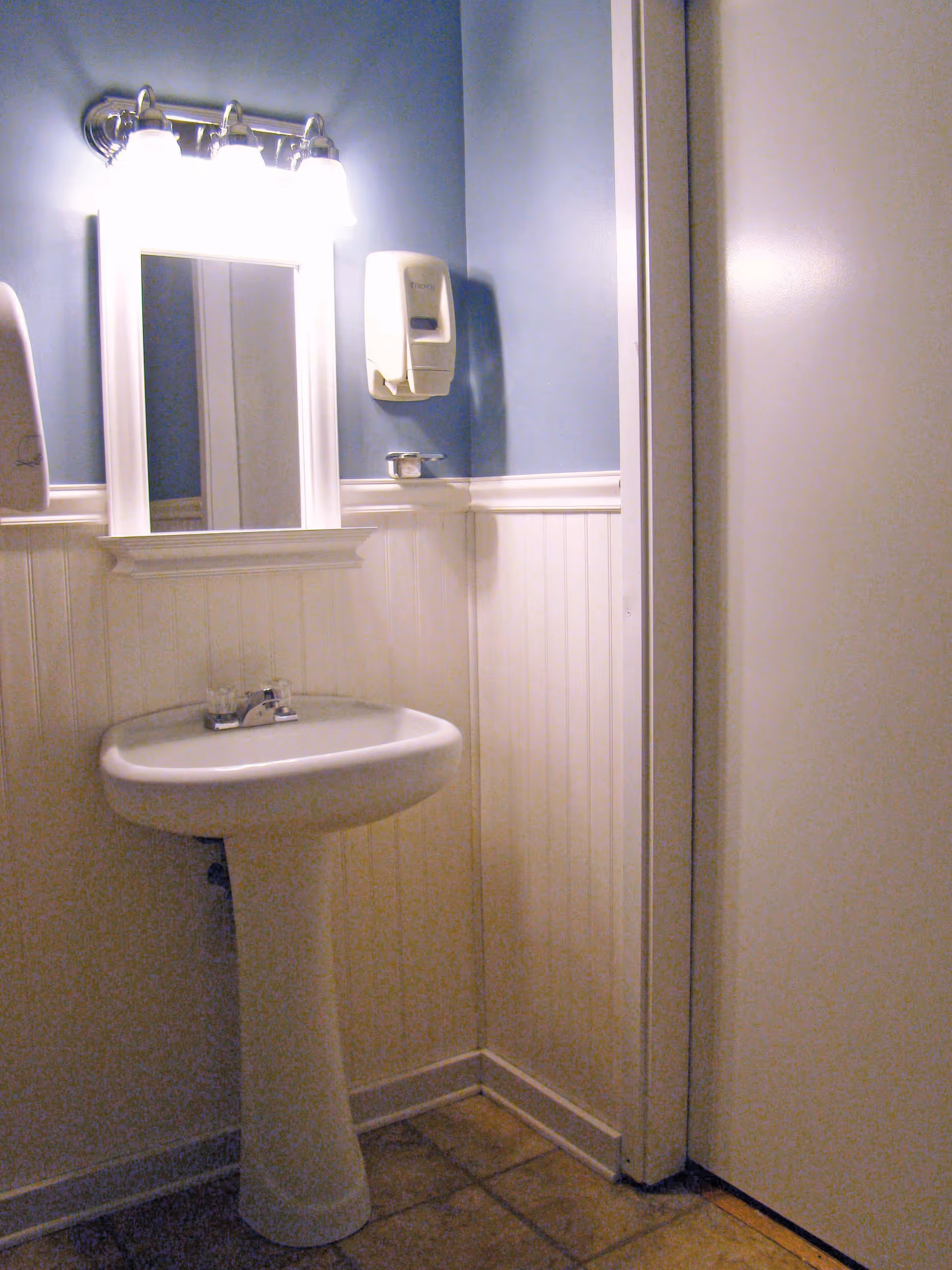 A small bathroom corner with a white pedestal sink, a rectangular mirror with a white frame above it, and a wall-mounted soap dispenser. The walls are painted blue on the upper half and white wainscoting on the lower half. There is a light fixture with three bulbs above the mirror and a closed white door to the right.