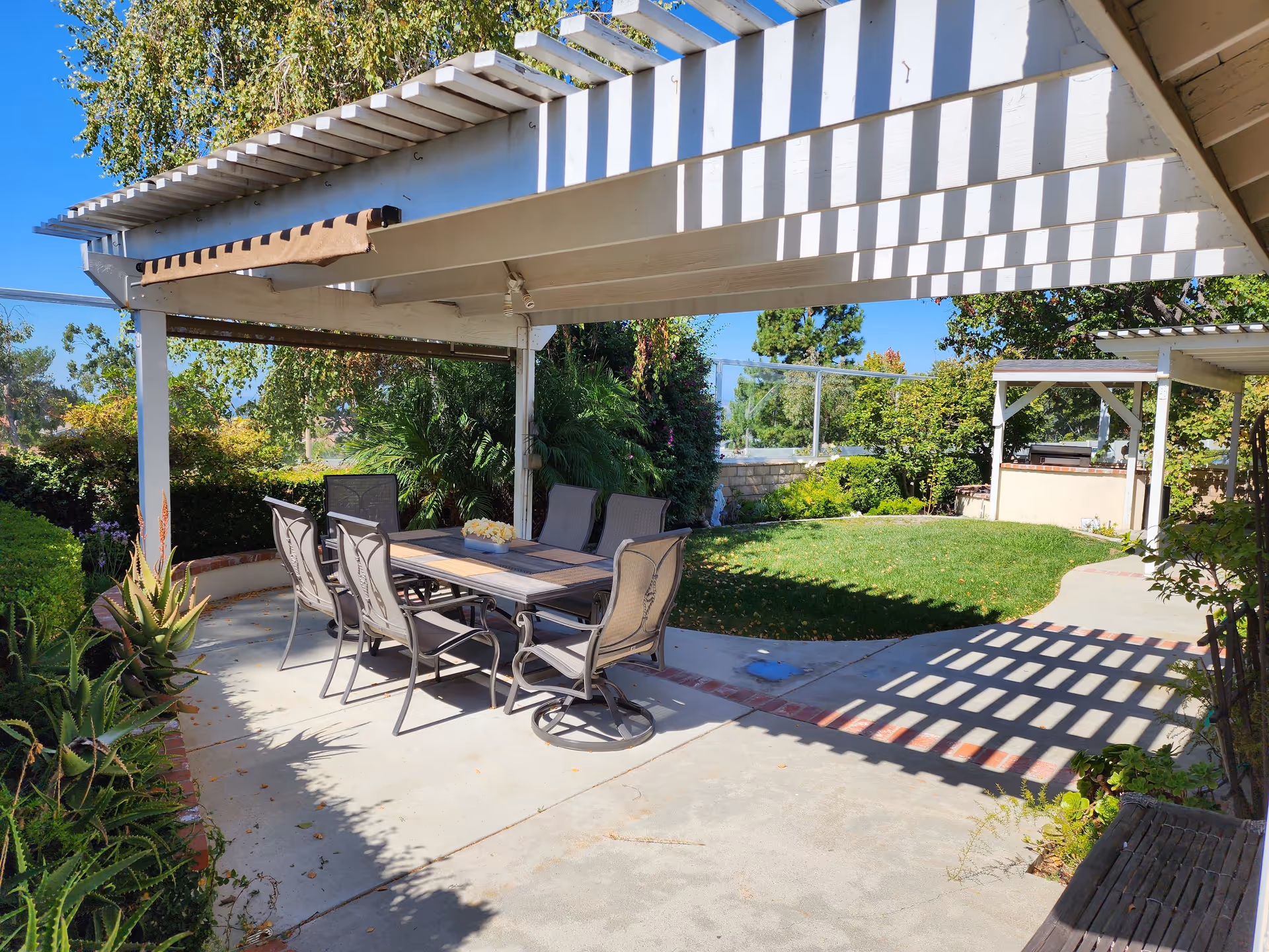 Outdoor patio area with a white pergola casting shadows on a concrete floor. There is a rectangular table with six chairs around it, surrounded by green plants and trees. In the background, there is a grassy lawn and a small structure with a roof, possibly a barbecue or outdoor kitchen area, under a clear blue sky.