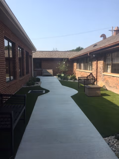 Outdoor courtyard area between two brick buildings with a concrete walkway, green artificial grass, benches, and a small table under clear sky.
