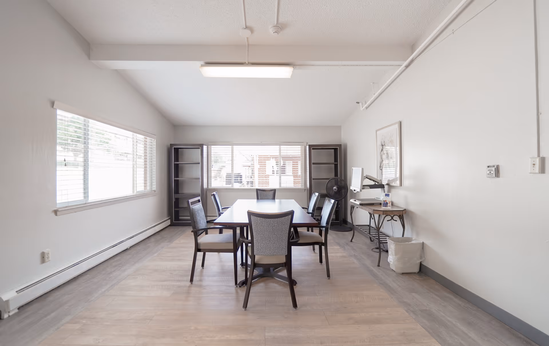 A bright, minimalist room with a rectangular table surrounded by six chairs. The room has large windows on two walls letting in natural light, two empty black bookshelves, a standing fan, a small table with a computer monitor, and a trash bin. The walls and ceiling are white, and the floor is light wood.