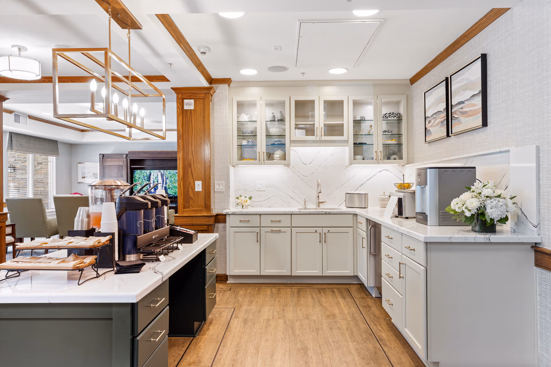 A modern kitchen area with white cabinets, marble countertops, and a wooden floor. The kitchen features glass-front upper cabinets displaying dishes, a sink, a coffee machine, a toaster, and a flower arrangement on the counter. There is a large island with coffee dispensers and cups, and a dining area with chairs and a window in the background.