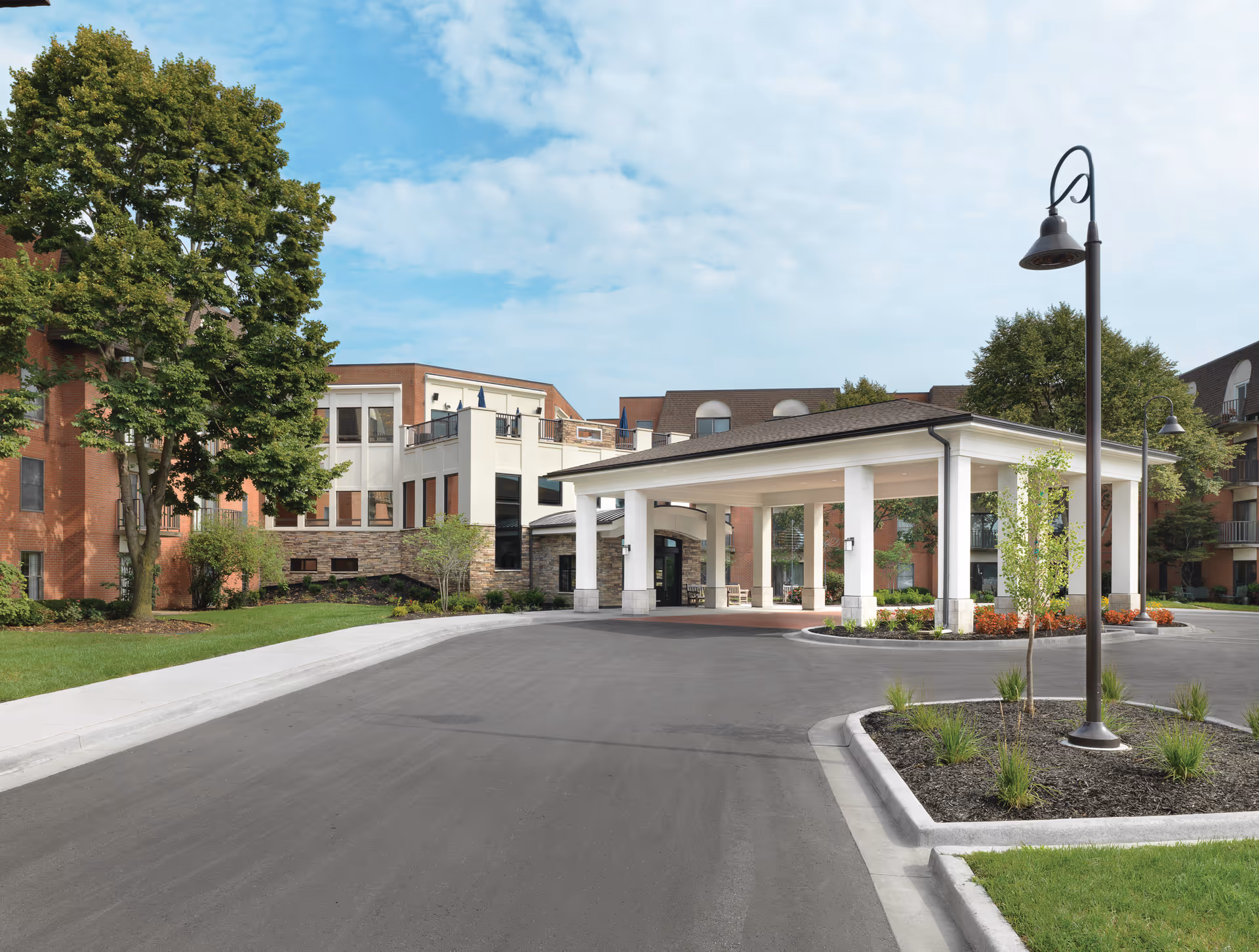 Exterior view of Kingswood Senior Living Community showing a covered entrance with white pillars, a paved driveway, landscaped areas with small trees and shrubs, and a multi-story brick building in the background under a partly cloudy sky.