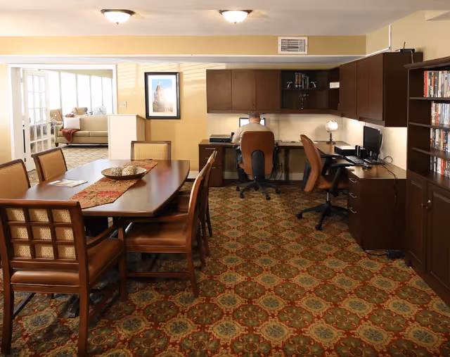 A room with a patterned carpet featuring a wooden table with six chairs on the left side and a workspace area on the right side with a man sitting on an office chair working on a computer. The workspace has multiple desks, chairs, shelves with books, and a small globe. In the background, there is a doorway leading to a living room area with a sofa and windows letting in natural light.