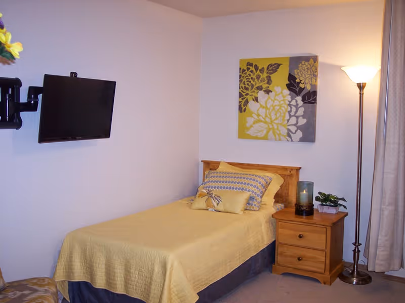 A small bedroom with a single bed covered in a yellow bedspread and multiple pillows. Next to the bed is a wooden nightstand with a candle and a small plant. A floor lamp stands beside the nightstand, and a flat-screen TV is mounted on the wall opposite the bed. A decorative floral painting hangs above the bed, and beige curtains cover a window on the right side.
