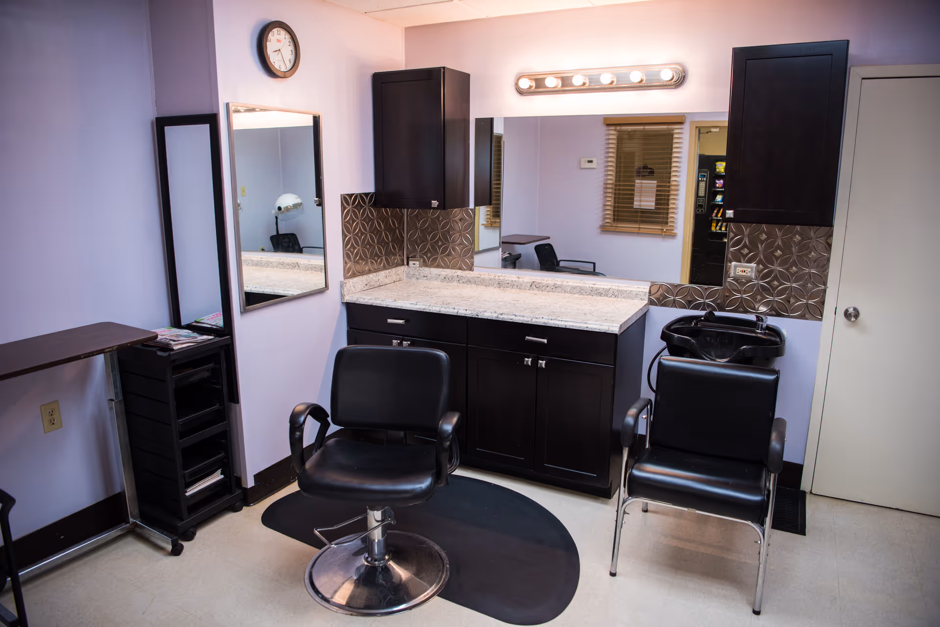 A salon-style interior with two styling chairs, a countertop with mirror and cabinets, and a wash basin.