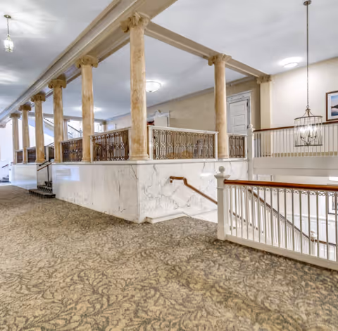 Interior view of a senior living facility hallway with ornate columns, marble half-walls, carpeted floor, and a staircase with white railings. The ceiling has recessed lighting and a hanging light fixture, with a framed picture on the wall.