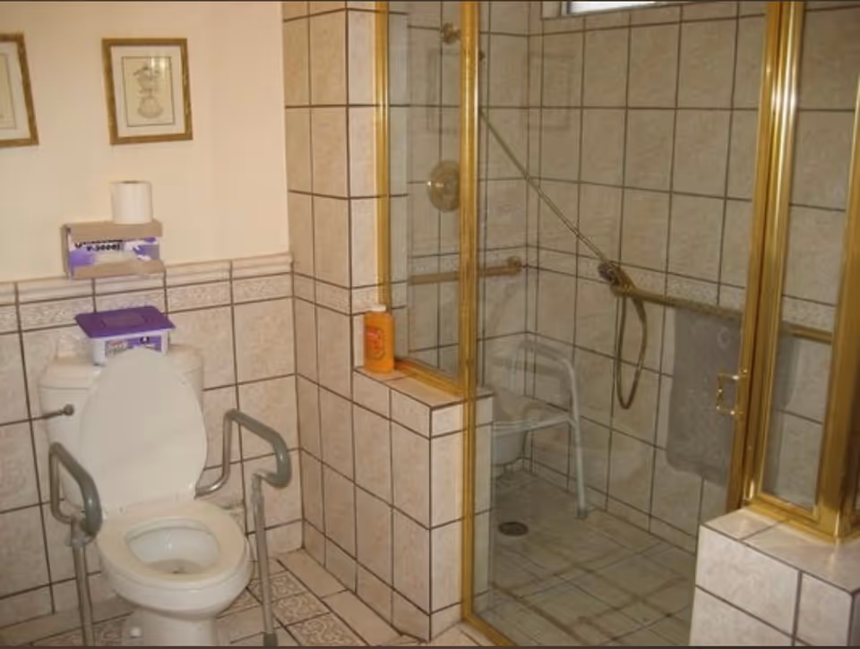 A bathroom with beige tiled walls and floor, featuring a toilet with safety rails, a roll of toilet paper on a holder above the tank, and a walk-in shower with a glass door and gold trim. Inside the shower is a white shower chair and a handheld showerhead. There are two framed pictures on the wall above the toilet.