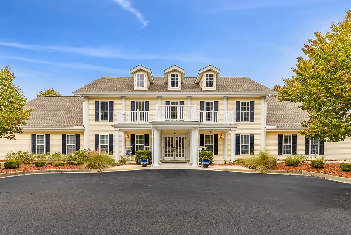 Front exterior view of a two-story senior living facility building with beige siding, black shutters, and a gray shingled roof. The entrance features white columns, a covered porch, and a balcony above. There are two potted plants flanking the entrance and landscaped bushes and trees on either side.