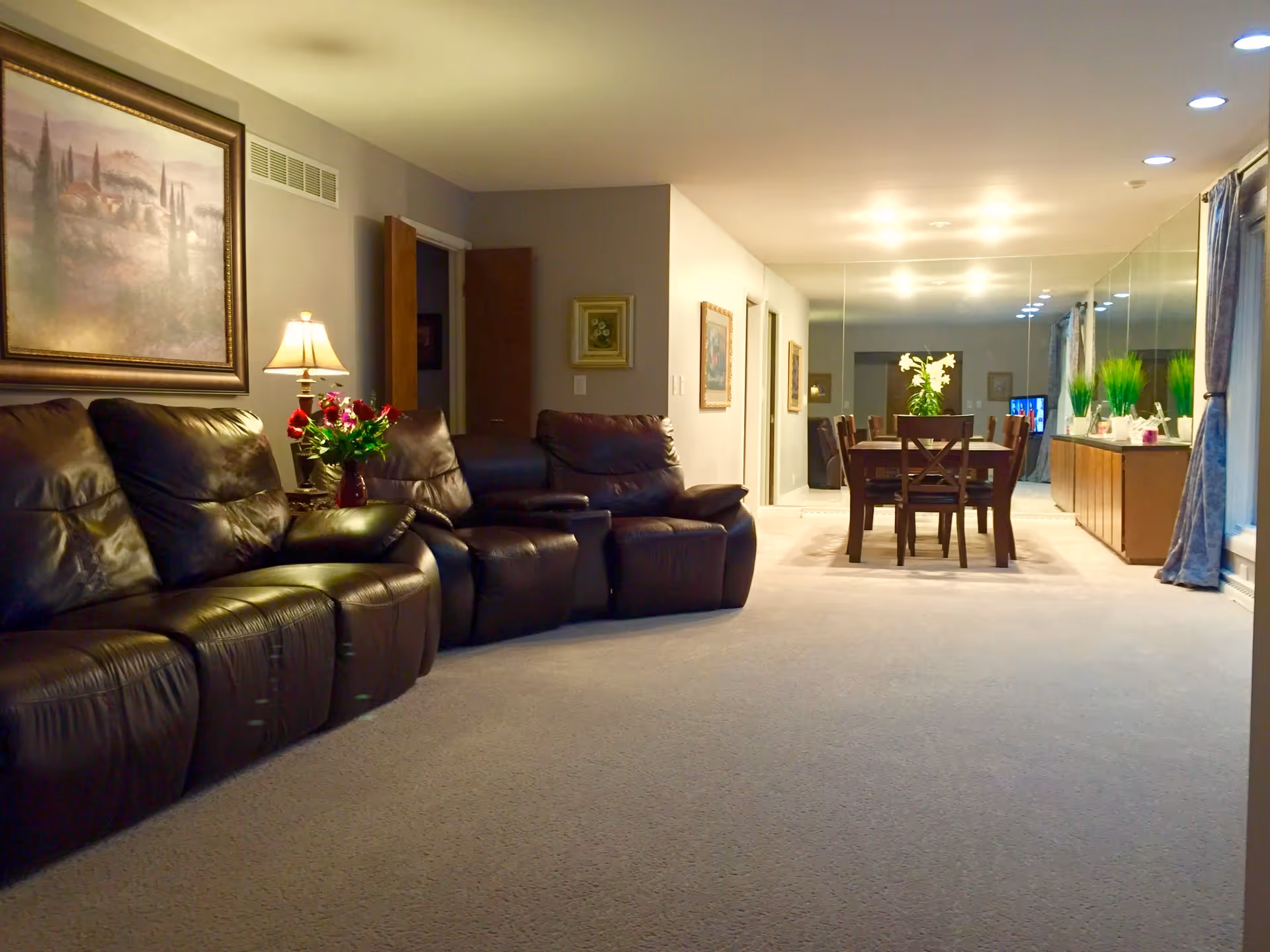 A cozy living and dining area featuring a row of dark brown leather recliners on the left, a side table with a lamp and a vase of flowers, and a dining table with chairs in the background. The far wall is covered with large mirrors reflecting the room, and there are decorative plants and framed artwork on the walls.