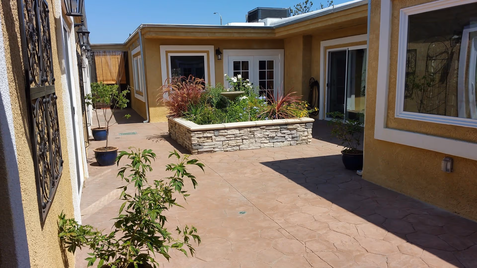 Outdoor courtyard area with a raised stone planter filled with various plants and flowers, surrounded by a paved walkway. The courtyard is enclosed by yellow stucco walls with white trim, windows, and glass doors. Several potted plants are placed along the walkway.