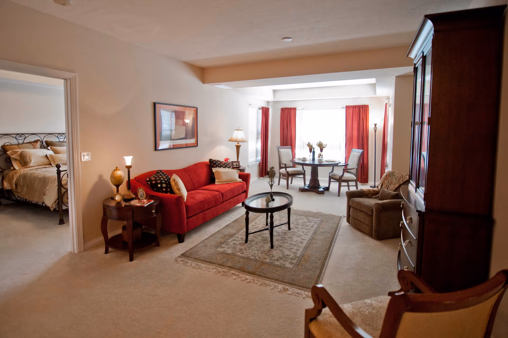 A cozy living room with a red sofa, a round glass coffee table on a patterned rug, a wooden side table with lamps, and a wooden cabinet. In the background, there is a dining area with a round table and four chairs near a large window with red curtains. To the left, an open doorway reveals a bedroom with a bed and pillows.