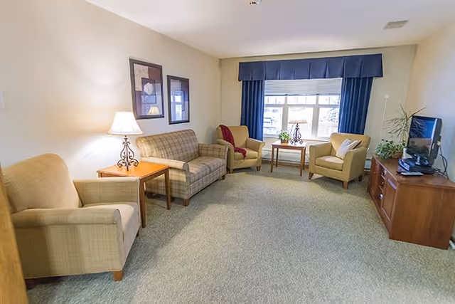 A cozy living room with beige carpet, a plaid sofa, three beige armchairs, a wooden side table with a lamp, a wooden TV stand with a flat-screen TV, and a window with blue curtains letting in natural light.