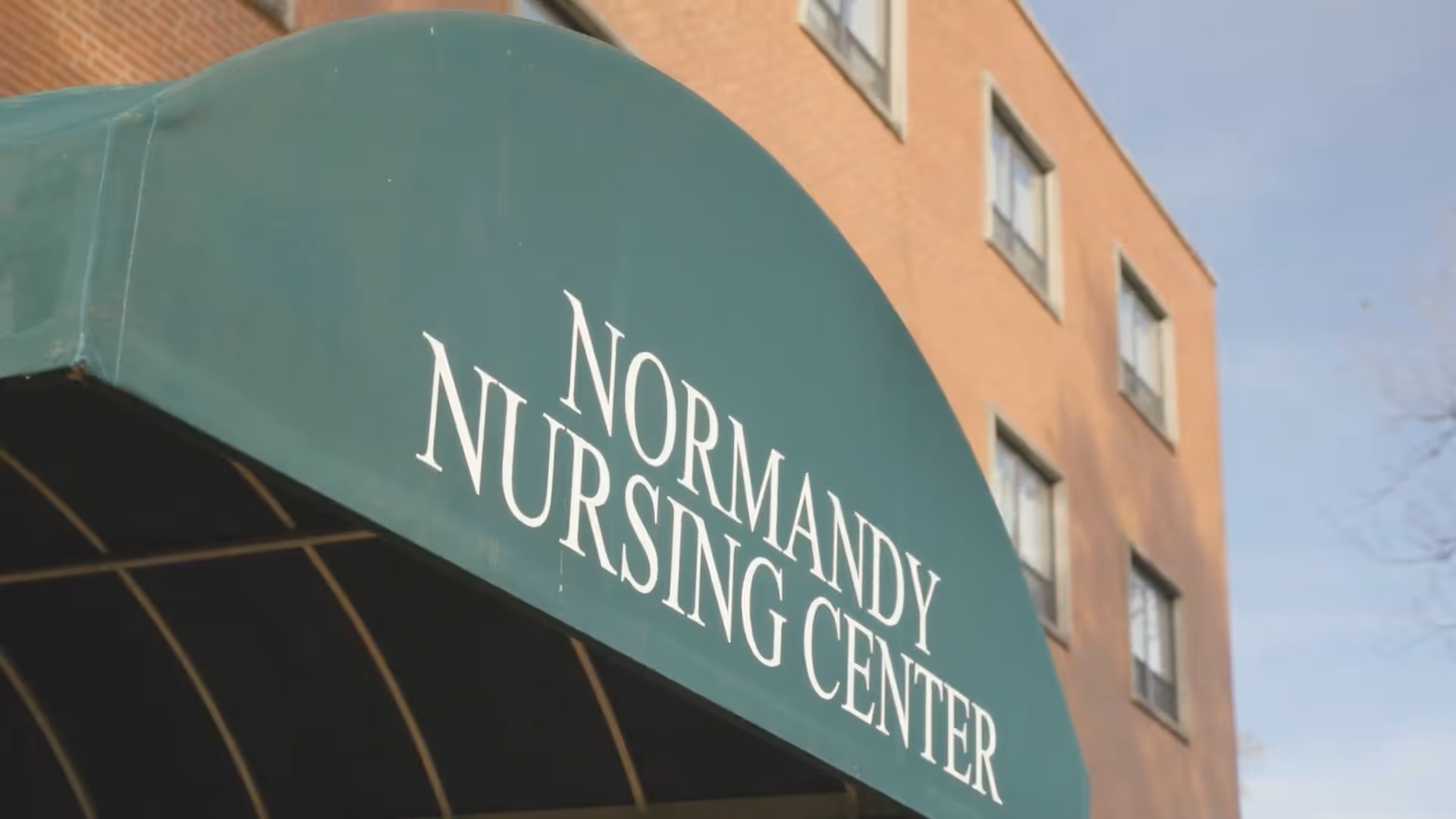 Green awning with white text reading 'Normandy Nursing Center' attached to a brick building with several windows under a clear sky.