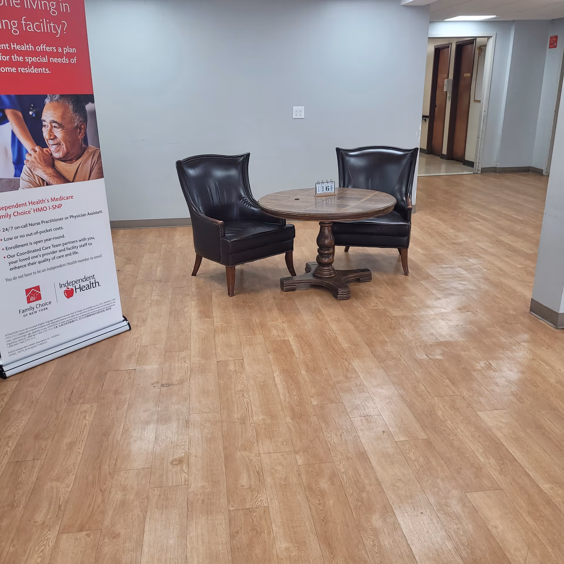 A small seating area in a nursing facility with two black leather chairs and a round wooden table between them. The table has a small calendar displaying the number 16. To the left, there is a standing banner with information about Independent Health and Family Choice Medicare. The floor is wood-patterned, and the walls are light gray. In the background, there is a hallway with doors.