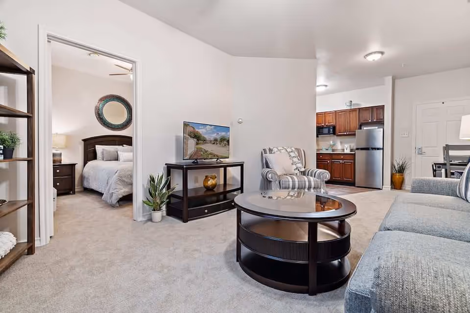 Furnished living room showing a sofa, round coffee table, TV on a stand, armchair, open kitchenette, and a bedroom visible through a doorway.