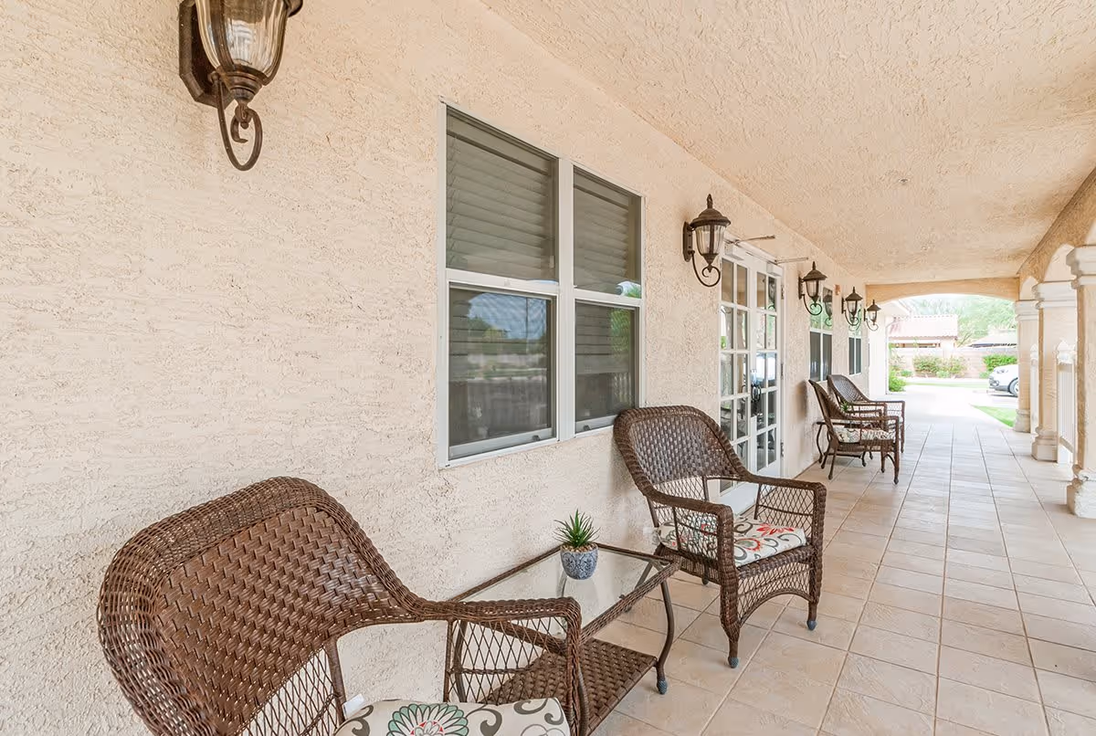 Covered outdoor patio area with beige textured walls and tiled floor, featuring several brown wicker chairs with patterned cushions and small glass-top tables. Wall-mounted lantern-style light fixtures are evenly spaced along the wall, and windows and glass doors are visible. The patio opens to a landscaped outdoor area with greenery and a parked car in the distance.