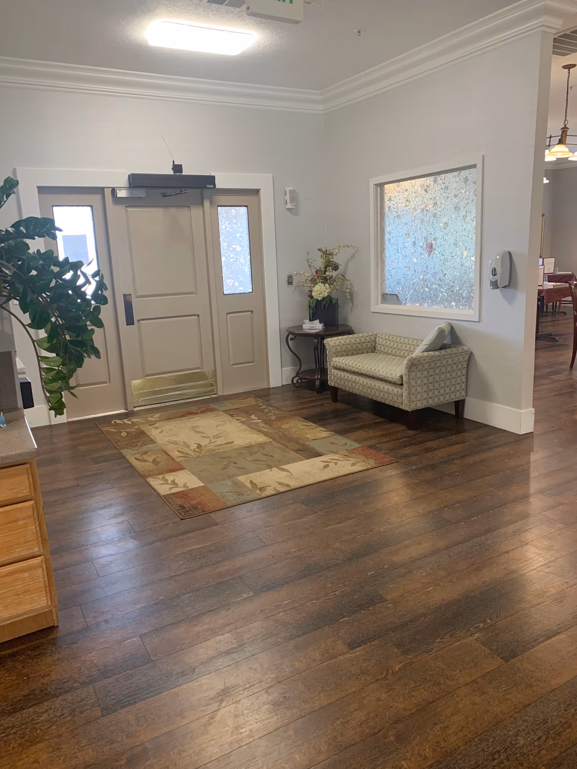 An interior entry foyer with double doors, a patterned area rug, wood flooring, a small upholstered bench and a side table with flowers.