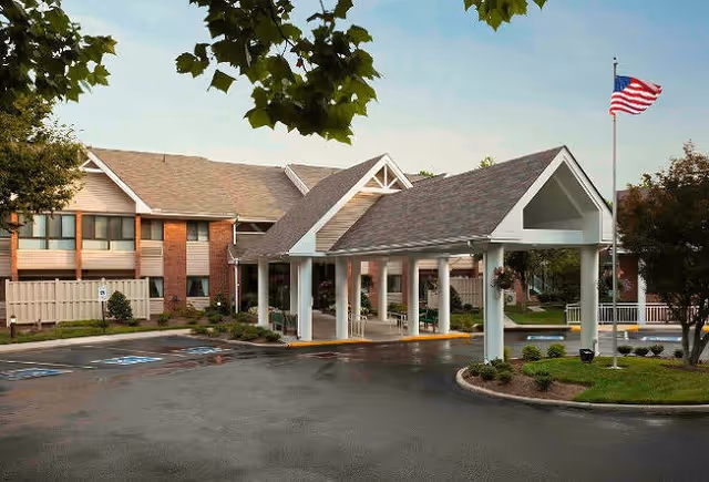Exterior view of a senior living facility with a covered entrance supported by white columns, a two-story building with beige siding and brick accents, an American flag on a flagpole, and landscaped greenery around the parking area.